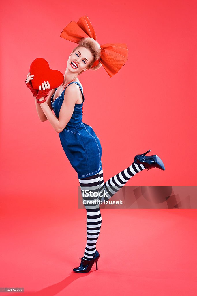 Happy woman with big heart Young blond beautiful woman wearing jeans dress, striped tights and big, red hair ribbon holding a big heart in her hands. Studio shot, red background. 20-24 Years Stock Photo