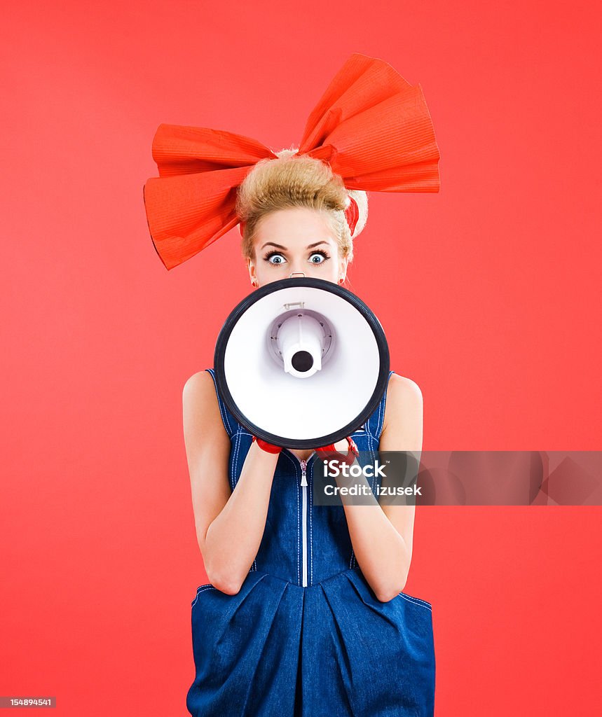 Woman with big hair ribbon shouting into megaphone Young blond beautiful woman wearing jeans dress and big, red hair ribbon shouting into megaphone. Standing against red background. Studio shot. Fashion Stock Photo