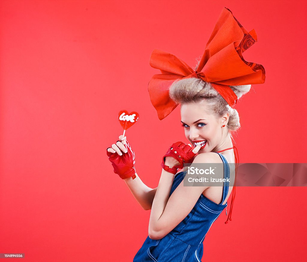 Happy woman with red lollipop Happy young blond beautiful woman wearing jeans dress and big and red hair ribbon, holding red lollipop with "love" word in her hand. Studio shot, red background. Fashion Stock Photo