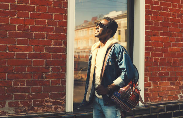 istock Stylish young african man wearing jeans jacket and backpack looking up at sunlight while walking on city street over brick wall background 1548973890