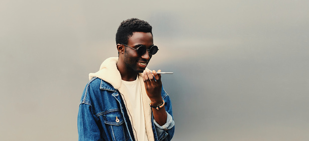 Portrait of modern young african man holding smartphone using voice command recorder, assistant or takes calling, looking at phone on gray background