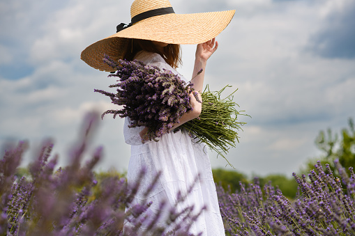 Girl in a white dress with a bouquet of fresh lavender on a lavender field. Wide-brimmed straw hat on the head.