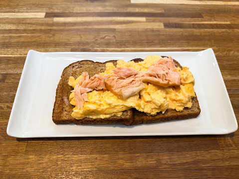 Scrambled Eggs. Fluffy and Buttery scrambled eggs on bread with microgreen radish and hollandaise sauce on white plate over white wooden background. Homemade breakfast or brunch meal. Top view.