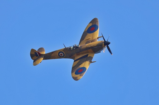 Spitfire Ww2 Battle of Britain fighter aircraft turning against a bright blue sky.  Sharp image highlighting the camouflage paintwork of the aircraft.  No identity markings on the aircraft. The photo was taken at Whittlebury Northamptonshire in the UK as the plane flew overhead.
