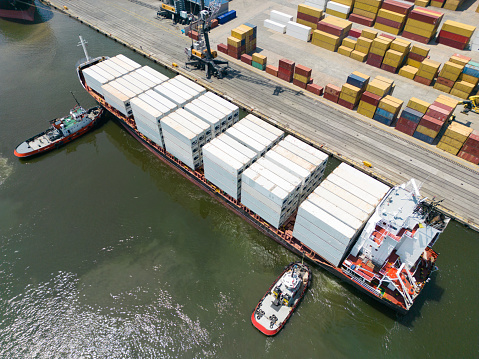 The tug accompanies the ship when entering the cargo port. \nAerial drone view above sea port. Cargo vessel is loading containers and bulk. Professional business logistics and transportation of cargo ship.