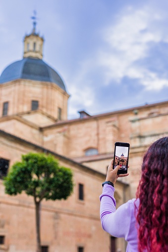 Lifestyle photo holding cellphone outdoors, in front of a historical monument in Castile and Leon, Plaza de las Angustias in Salamanca, spain. Famous church catholic religion building. ancien architecture in Europe with people