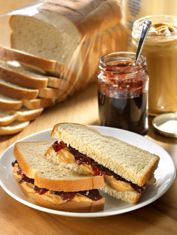Peanut Butter and Jam sandwich on whole grain white bread with peanut butter jar and jam jar in background with loaf of bread and bottle of milk.