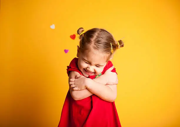 Color image of a little girl giving herself a big Valentine hug with hearts in the background on yellow. 