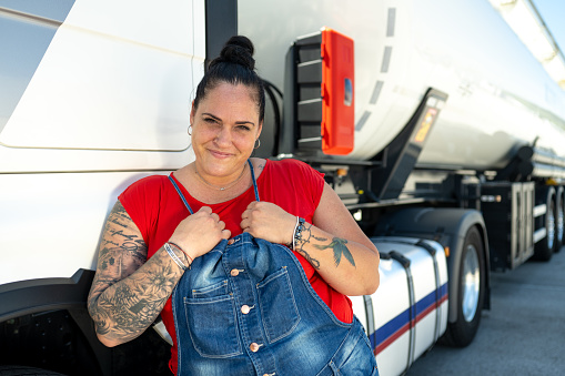 Close up of smiling heavy female truck driver leaning against a cabin door of a cistern looking at camera.