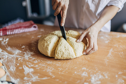 The woman cuts the dough into smaller pieces with a knife. She is preparing a pastry, there are ingredients on a wooden table. Unrecognizable person.