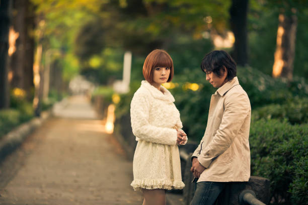 a young japanese couple in a public park appears to be getting to the end of their relationship, the guy is looking down, while girl looks at camera.