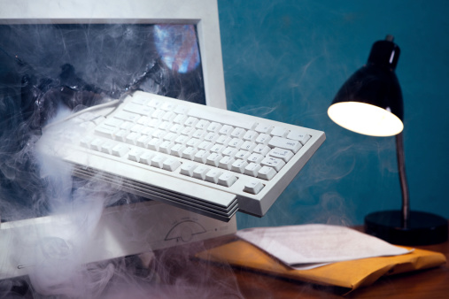 A destroyed computer monitor sitting on an office desk smokes through its broken glass screen, a keyboard slammed through the middle.