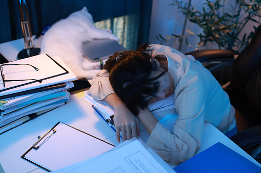 The woman's office was very tired and fell asleep at the workplace, her head lying on the table. A lot of paperwork. Tired employee, a stack of documents and papers. Sleepy tired alone girl dozing off at the table while working hard at late night.