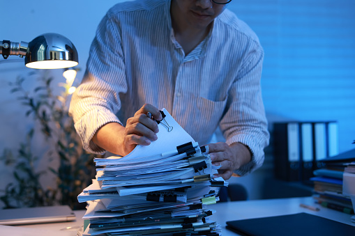 Asian young businessman hands working on Stacks of paperwork on his desk at home, work from home concept, business report, piles of unfinished documents, closeup view.