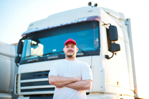 homem motorista de boné vermelho parado na frente de caminhão branco no parque de caminhões com os braços cruzados - semi truck driver pride white - fotografias e filmes do acervo