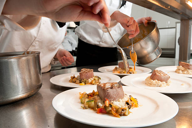 close up on chefs preparing white plates with grilled tuna, risotto and ratatouille in commercial kitchen, selective focus on the background