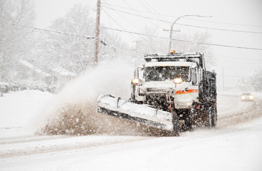 A town highway department snowplow dump truck is plowing snow and spreading road salt to clear a suburban residential district street during an extreme weather winter blizzard snow storm.