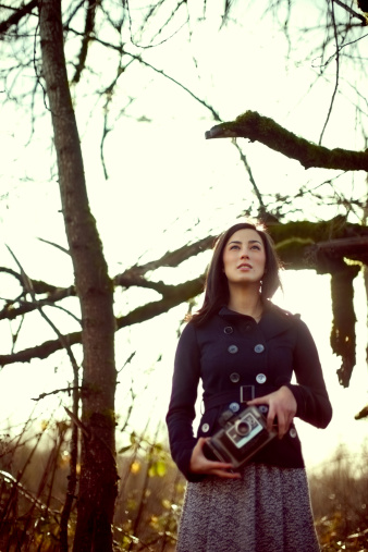 A beautiful Asian American woman stands in a nature area wearing a pea coat, the sun shining warm light throught the trees behind her as it sets.  Vertical with copy space.