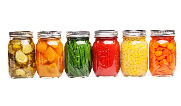 Row of canning jars featuring six different canned vegetables and fruits lined up and preserved in glass storage. The food varieties that are sauced, pickled, or sliced include carrots, green beans, tomatoes, corn, dill pickles, and sweet potatoes. Horizontal straight-on view, cut out and isolated on a white background.