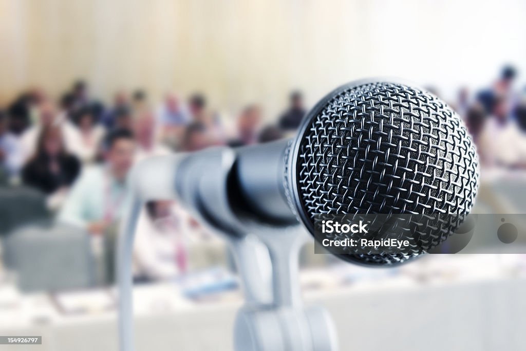 Close up microphone; seminar attendees in background A vocal microphone stands ready against a background of an out-of-focus and unrecognizable audience at a seminar or meeting.  Convention Center Stock Photo