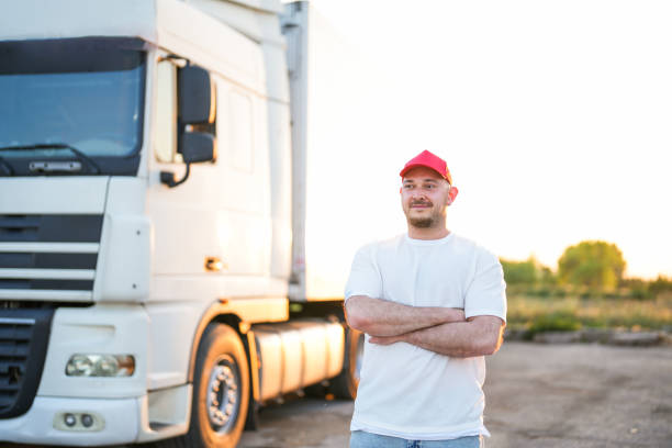 homem motorista de boné vermelho parado na frente de caminhão branco no parque de caminhões com os braços cruzados - semi truck driver pride white - fotografias e filmes do acervo