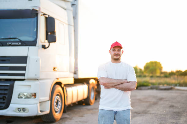 homem motorista de boné vermelho parado na frente de caminhão branco no parque de caminhões com os braços cruzados - semi truck driver pride white - fotografias e filmes do acervo