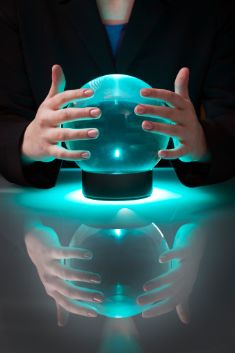 Subject: Vertical view of an unidentifiable person seated by a reflective table with their hands around a crystal ball glowing with an eerie blue light, rubbing it to predict the future.