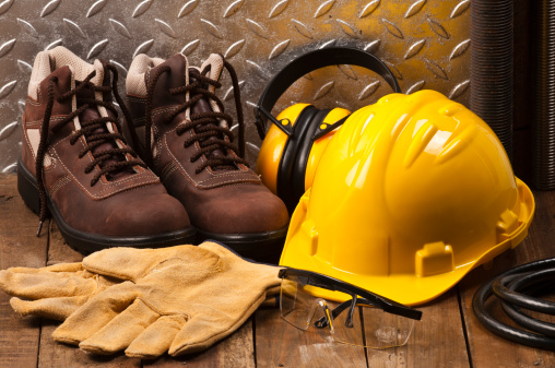  Personal Protective Workwear Shoot on Work Location. The Safety items are placed on rustic wood and includes a Yellow Hard Hat, Gloves, Steel Toe Shoes, Ear Muff and Goggles. A silver diamondplate in the background. Predominant colors are yellow and brown