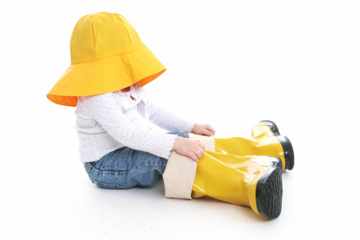 Profile of unrecognizable little girl in rain clothes with adult oversized yellow hat and rain boots.  Full body showing on white background. Part of an on-going series.