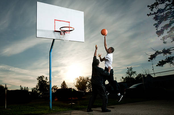 a basketball hoop attached to a metal mesh backboard, mounted on a vertical support. The hoop has a red rim, and the net is significantly frayed with several loose strands. Bare tree branches are visible in the dark background, indicating a nighttime setting.