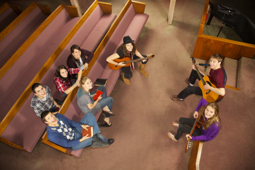 Eight teen friends play guitar and worship together in church setting as seen from high angle view