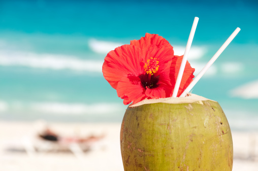 A tropical coconut cocktail decorated with a colorful bloom on a Caribbean beach. The beach and waves in background are out of focus.