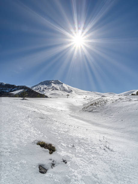 A dramatic sunset image featuring the silhouette of a mountaineer with a heavy backpack standing on a snowy mountain summit. The human figure appears small on the right, facing an endless expanse of colourful clouds and a glowing sun, with the stratosphere line clearly visible in the sky. This powerful scene from Monte Vettore highest peak of Sibillini National Park evokes themes of adventure, perseverance, and nature's majesty.