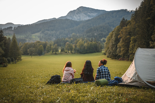 Three young women sitting on a slope and enjoying the view of the hills, their tent behind them. Full length shot, back view.