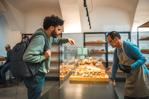 Middle Eastern male customer standing in line in a small artisan bakery. Buying freshly baked goods. Cheerful Asian male shop assistant handing them their orders.