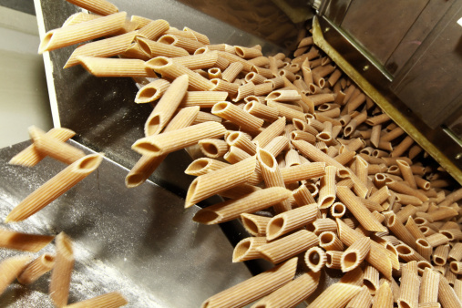 Whole wheat rigatoni falling from an industrial pasta maker, flour specks flying around. Other images in: