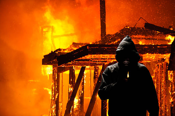Hooded person holding a lighter in front of burning house stock photo