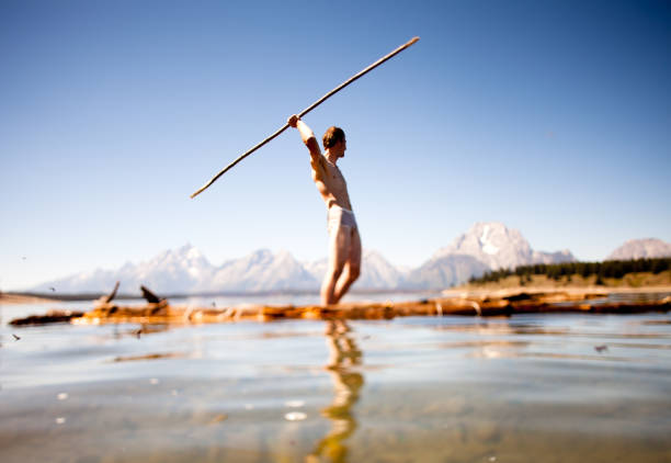 Boy standing on lakeside with a long stick, playing near the water in nature on a summer day
