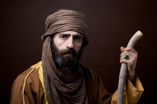 Middle eastern man with headscarf in traditional headscarf and clothing.the model is 40 years old.He is holding a wooden rod, wearing a brown headscarf and yellowish brown frock.There is a ring with green stone in his finger.The photo was shot in studio with a full frame DSLR camera in horizontal framing.