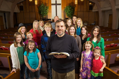 A friendly,happy church with their pastor smiling and facing the camera while inside a traditional church sanctuary and cross on wall in background,cross,pews,indoors,group,large group of people,group shot,facing camera,expressing positivity, multi generational,men and women, boys and girls, cross in background,copy space