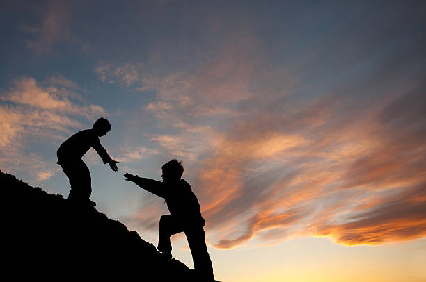 Lending A Helping Hand To a Brother In Need Concept image of two boys climbing a hill with one reaching out to help the other. Silhouette. Elementary age children. Side view. Teamwork concept. Additional themes in the image are: helping, friendship, siblings, brothers, lending a hand, assisting, caring, support, climbing, hiking, fitness, love, reaching out, neighborly, giving, stretching, gratitude, care, relationships, and family.  climbing up a hill stock pictures, royalty-free photos & images