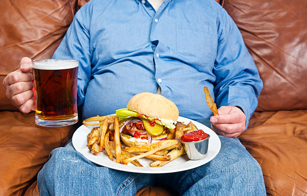 Obesity is a major cause of diabetes A photo of an overweight man sitting on an old couch with a very large unhealthy meal on his lap and a pint of beer in his hand. Obesity is a major cause of diabetes. unhealthy eating stock pictures, royalty-free photos & images
