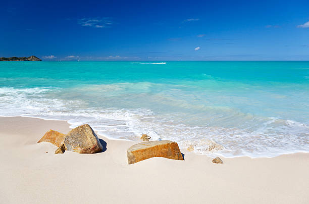 A clean white caribbean beach with deep blue sky and turquoise water, some stones in the foreground. Fort Beach in Antigua.
