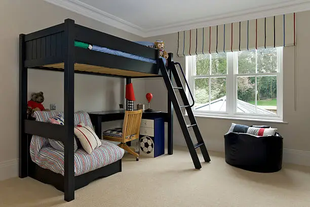 a young boy's bedroom in a new home. A large wooden framed bunk bed is located in the corner of the room adjacent to a large window overlooking the garden. A ladder leads up to the child's sleeping area which contains a mattress and duvet. Teddy bear's and toys together with a writing desk and drawers typify this as a boy's room. A large beanbag sits to the right.
