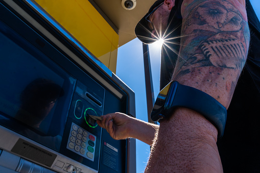 On a warm summer day, a man in his early 30s wears a black hat, shirt with arm tattoos, glasses, and colorful shorts. Engaged in using an outdoor ATM, he maintains a cheerful demeanor. Against the backdrop of blue skies, passing cars animate the scene, with a majestic church nearby and distant mountains adding to the picturesque view.