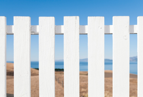 Close-up on a freshly painted wooden picket fence, with a scenic background of hills, ocean and summer sky.  Shallow depth of field, with focus on the fence.