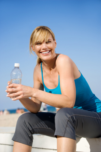 Attractive adult woman in sports clothing, holding water bottle ready to exercise on beach. She is beautiful and embracing a healthy lifestyle on a beautiful day.
