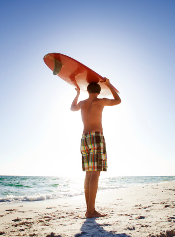 Teenage kid looking away while holding a red surf board above his head. Vertical shot.