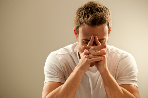 A head and shoulders image of a young Caucasian man with short brown hair dressed in a white t shirt leaning on his hands with fingers pointing towards his forehead, against a plain background. This could depict pain, depression, deep thought, or grieving and loss. With copyspace. 