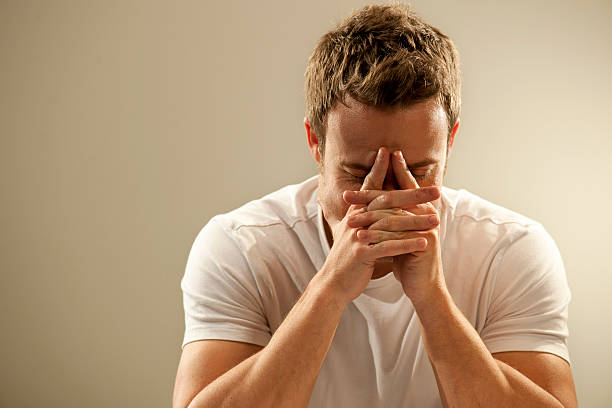 Young Man Thinking A head and shoulders image of a young Caucasian man with short brown hair dressed in a white t shirt leaning on his hands with fingers pointing towards his forehead, against a plain background. This could depict pain, depression, deep thought, or grieving and loss. With copyspace.  head in hands stock pictures, royalty-free photos & images